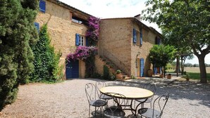 Un bâtiment rustique en pierre, doté d’une cour en gravier, d’une table ronde entourée de chaises et d’un escalier menant à un deuxième étage.