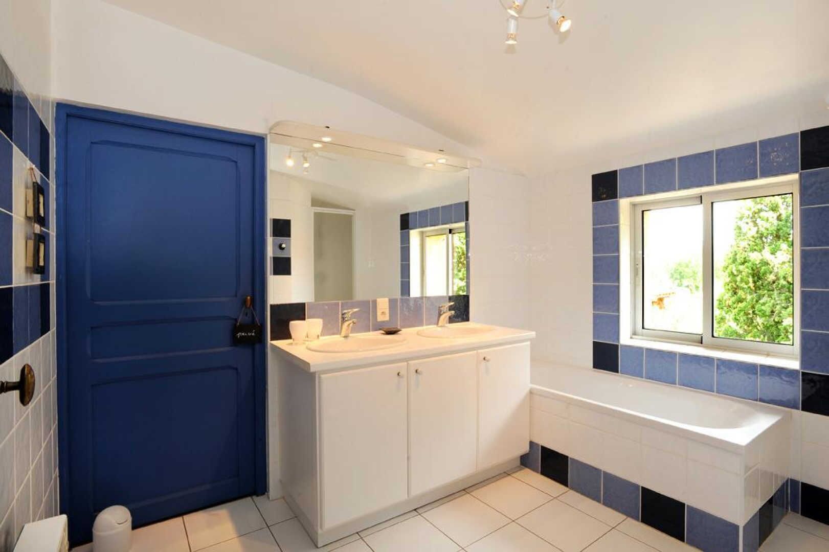 Une salle de bain avec une porte bleue, des meubles blancs, un lavabo et une fenêtre donnant sur un paysage verdoyant.