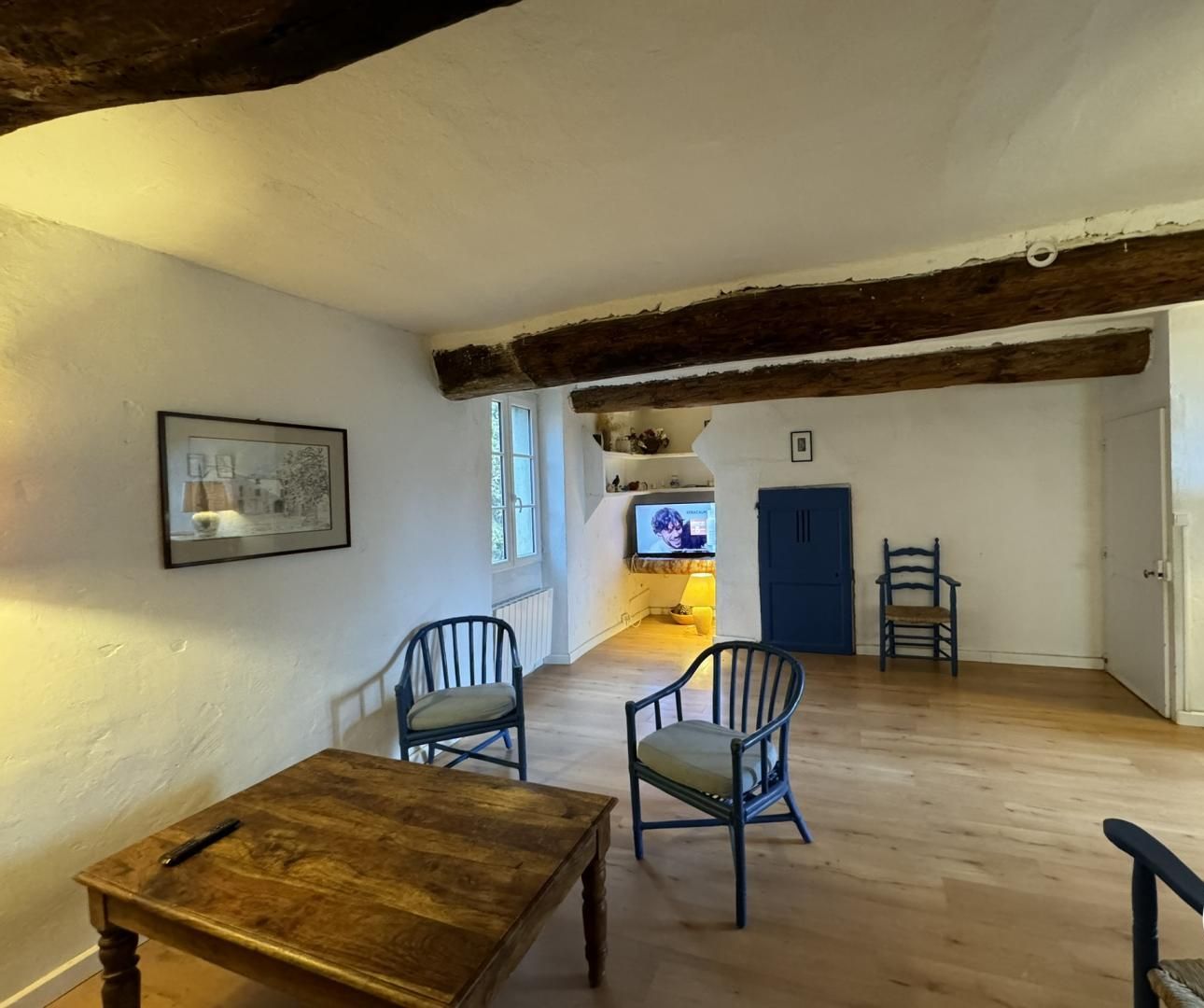 Una habitación amplia con una mesa de madera y dos sillas, un televisor al fondo y una puerta azul.