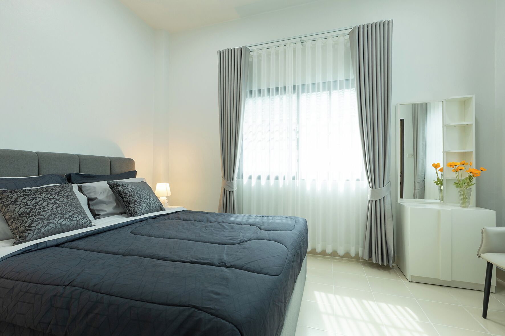 A modern bedroom with a large bed, a bedside table with a lamp, a dresser, and a mirror.