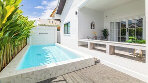 A swimming pool with a tiled deck, surrounded by a white building with large windows and a covered patio area.