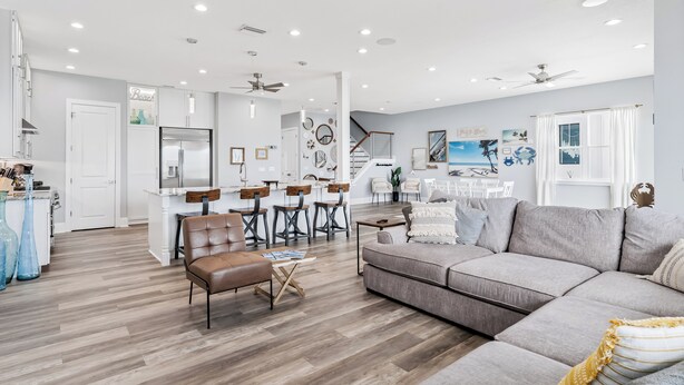 Un vaste salon avec un canapé gris, un fauteuil en cuir marron et une table basse. On y trouve également un coin repas avec une table et des chaises blanches. La pièce est dotée d’un parquet et de murs blancs.