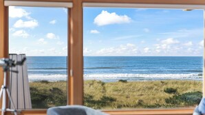 Una habitación con un ventanal grande con vista a una playa y dunas cubiertas de hierba.