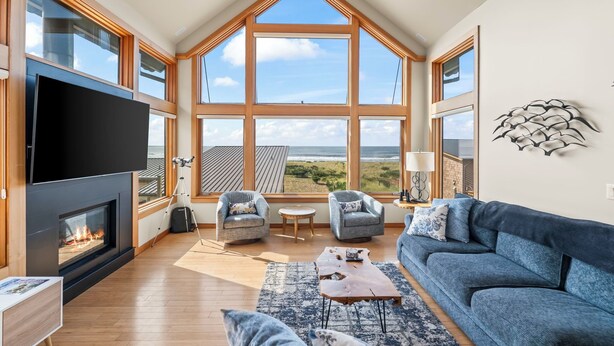 Un salón amplio con televisor grande, chimenea y vista al océano a través de ventanas grandes.