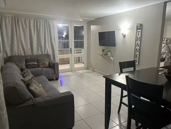 A living room with a sectional sofa, a dining table with chairs, a flat-screen TV mounted on the wall, and a sliding glass door leading to an outdoor area.