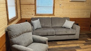 Une pièce chaleureuse avec un canapé et un fauteuil gris, des murs en bois et une fenêtre équipée de stores.