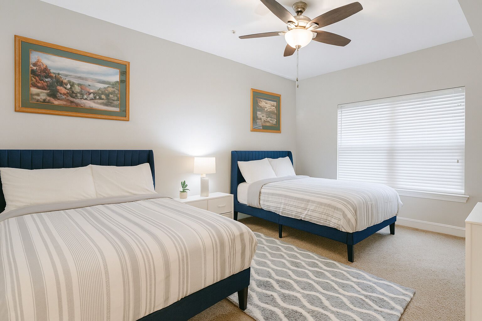 A bedroom with two beds, a ceiling fan, a window with blinds, and two framed pictures on the wall.