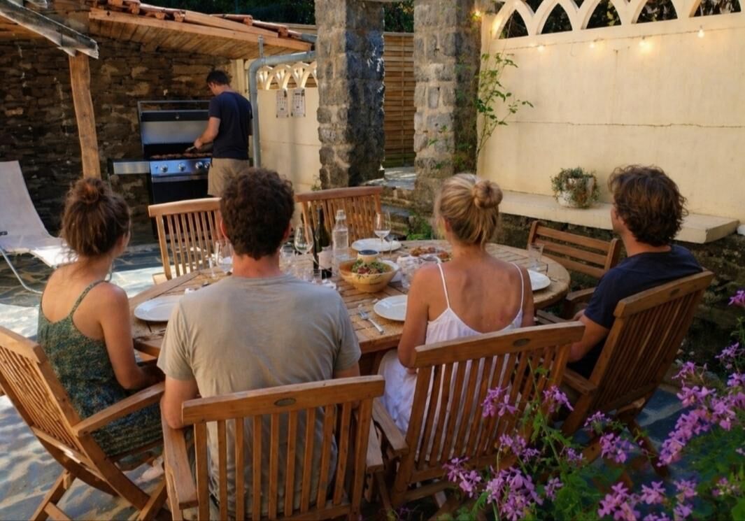 Un groupe de personnes est assis à une table en bois, à l’extérieur, devant un mur de pierre, avec un barbecue en arrière-plan.