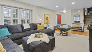 A modern living room with a grey sectional sofa, a blue armchair, and a white fluffy ottoman. There is a red door and a staircase in the background. - Spacious Family Retreat: Central/Hot Tub/Pool Table/Fire Pit/Kids Toys/3KingBeds (Arvada)