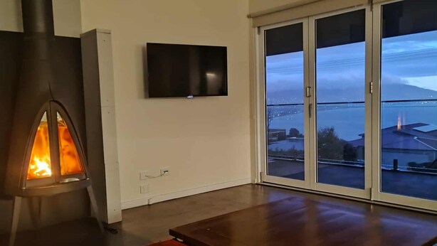 Uma sala de estar com uma lareira a lenha, uma janela grande com vista para a cidade e uma televisão de tela plana fixada na parede.