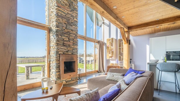 A spacious living area with a stone fireplace, large windows, a modern kitchen, and a view of the outdoors. - Modern 6-person Villa With Private Hot Tub - Near Beach & Nature (De Koog)