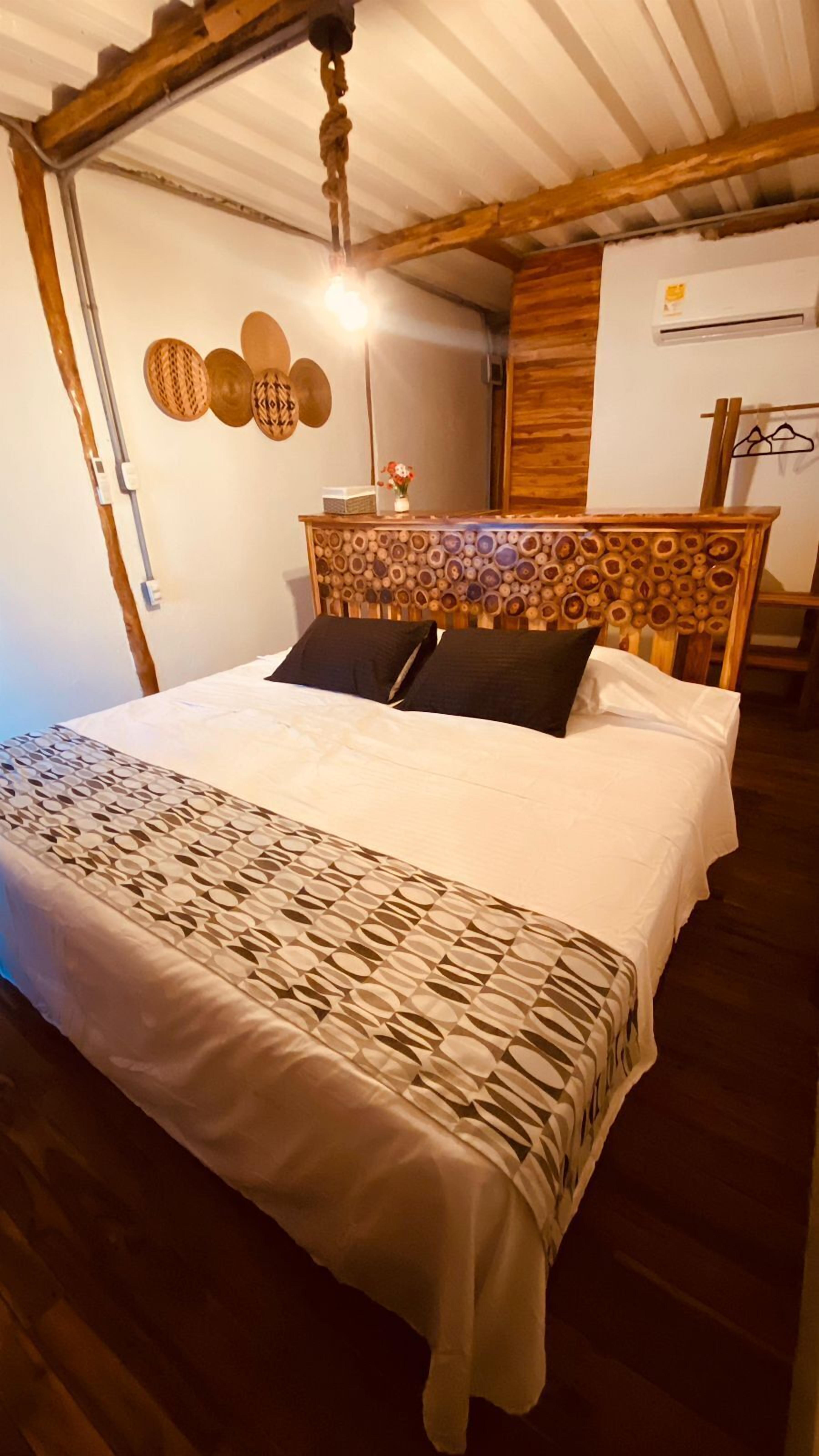 Quarto com cabeceira de madeira, cama com roupa de cama estampada, luminária pendente e uma pequena mesa com um vaso.