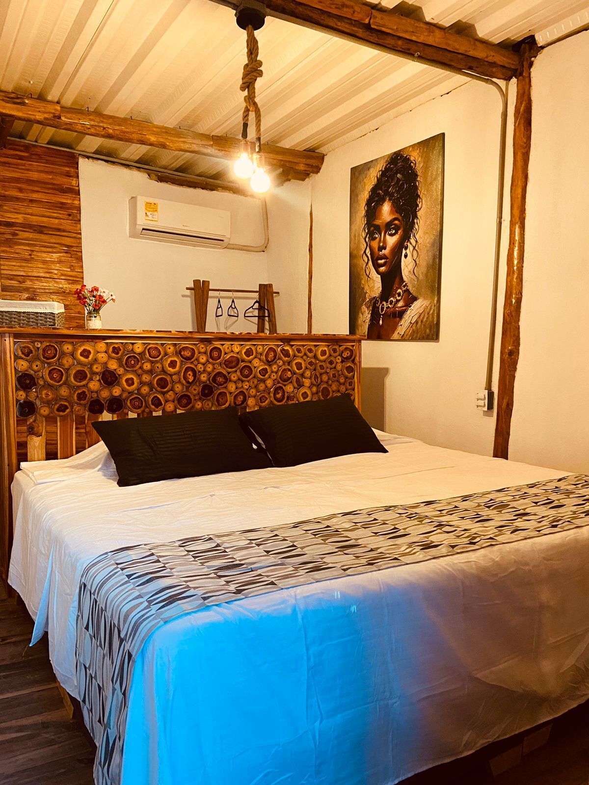 A bedroom with a wooden headboard, a bed with a patterned bedspread, a painting on the wall, and a hanging light fixture.