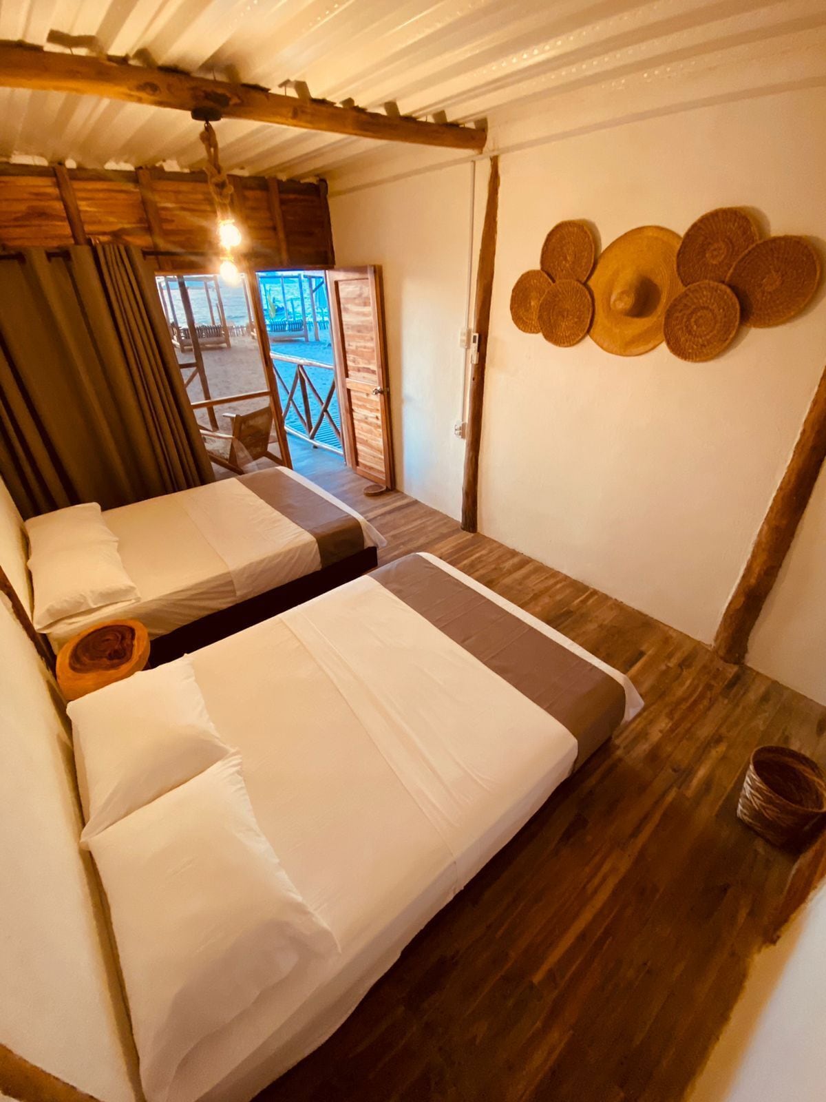 Habitación con dos camas, piso y techo de madera, y vista a la playa por una puerta abierta.