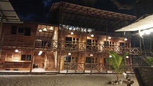 A wooden building with multiple balconies and large windows, surrounded by a sandy area with a palm tree. - Rocha Beach Club (Cartagena)