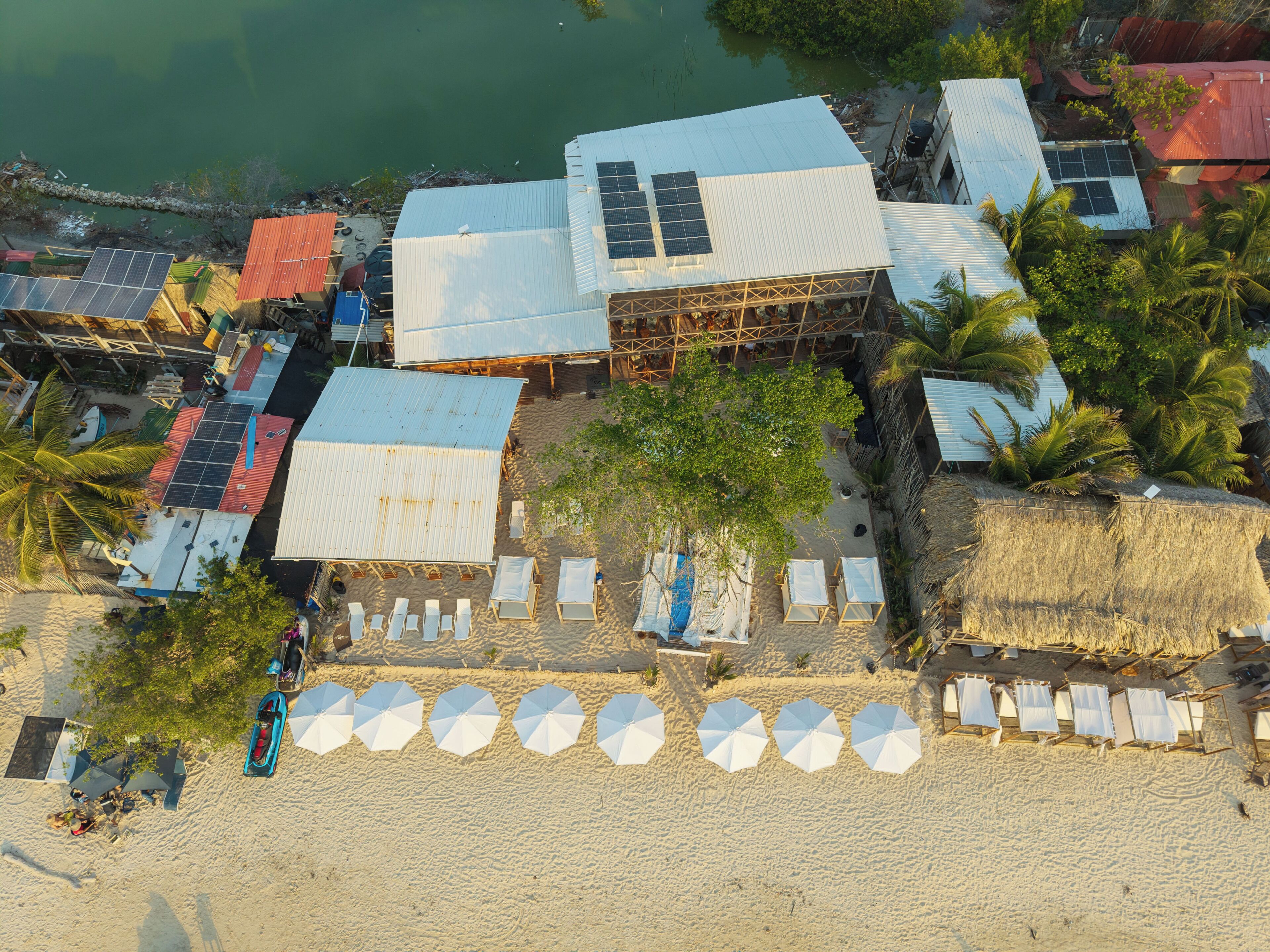 Una propiedad frente a la playa con varias habitaciones, algunas con paneles solares, y una hilera de sombrillas de playa.