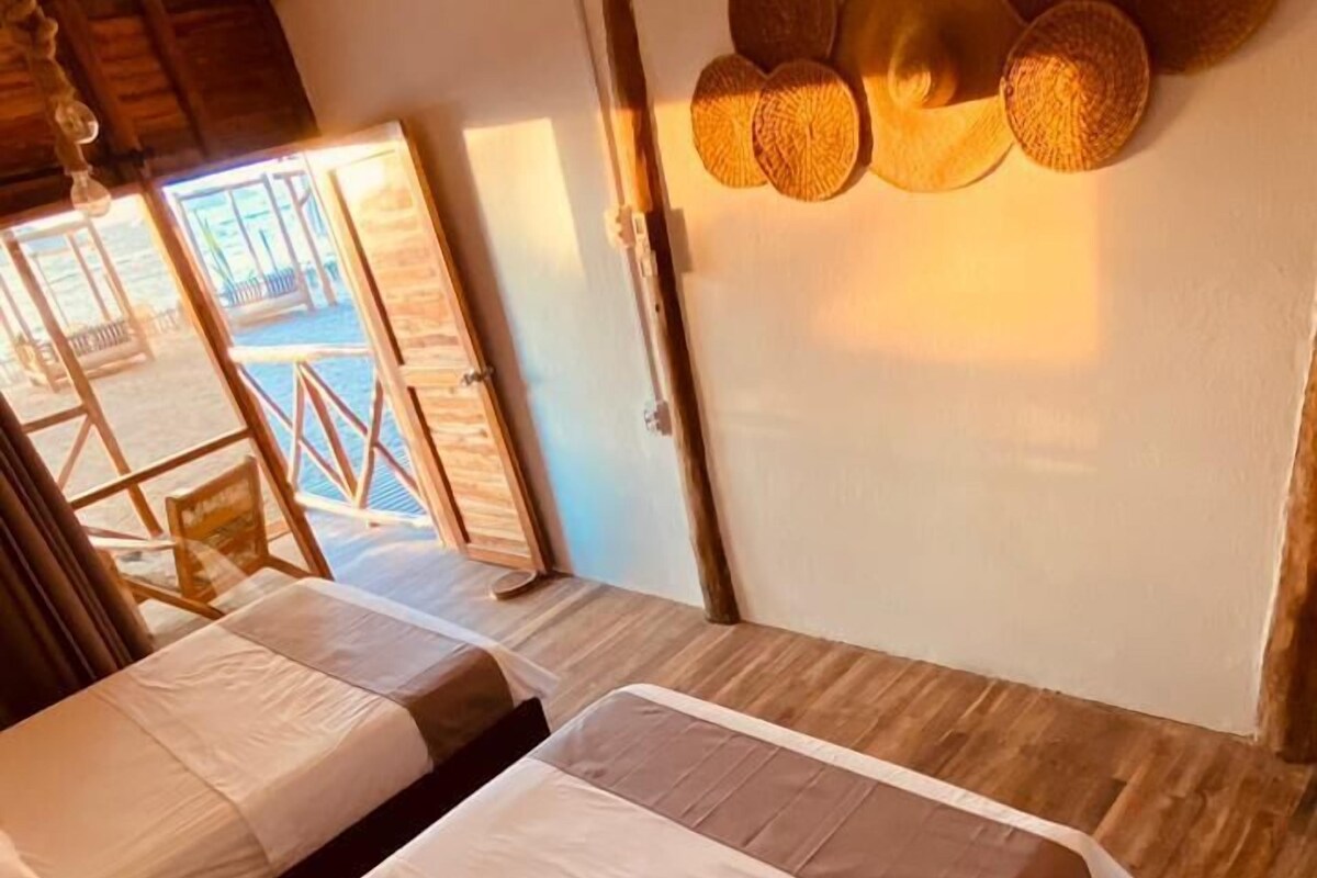 A room with two beds, wooden flooring, a wall-mounted light fixture, and a view of an outdoor area with a wooden deck and a pool.
