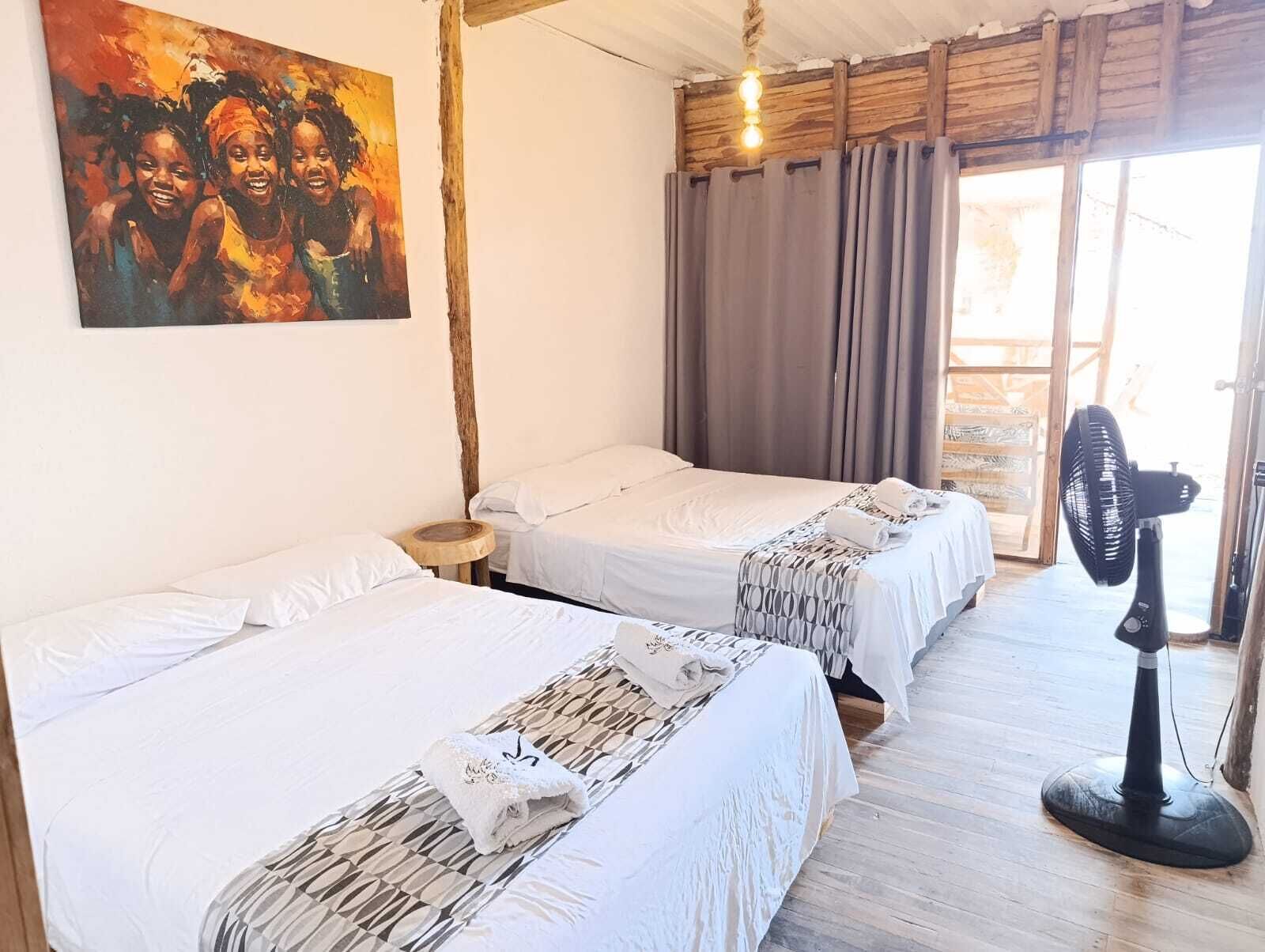 A room with two beds, a painting on the wall, a fan, and a window with curtains.