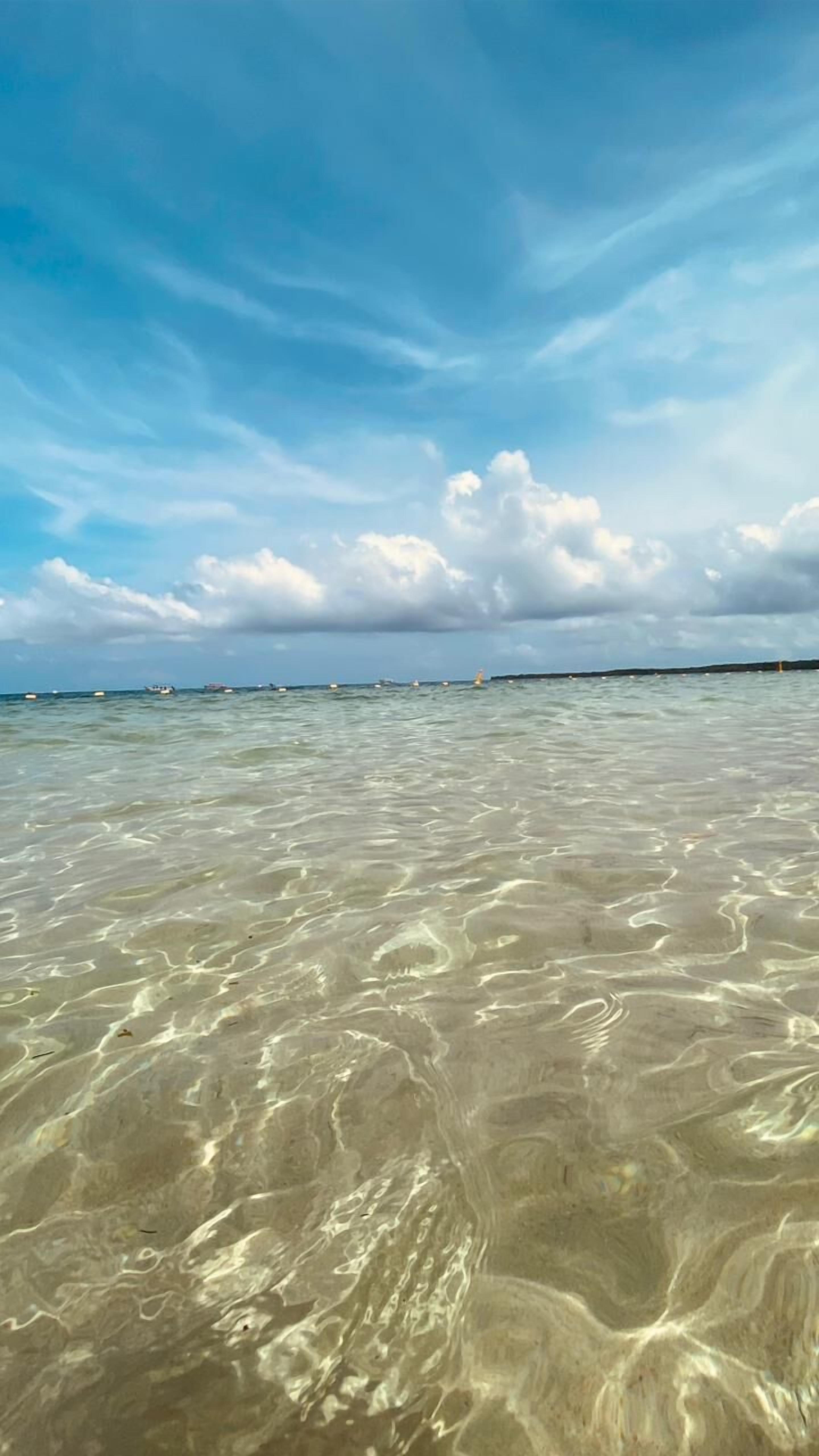A clear view of a beach with shallow water, distant boats, and a cloudy sky.