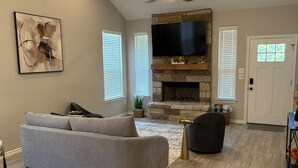 A living room with a gray sofa, a fireplace, a ceiling fan, and a flat-screen TV mounted above the fireplace.