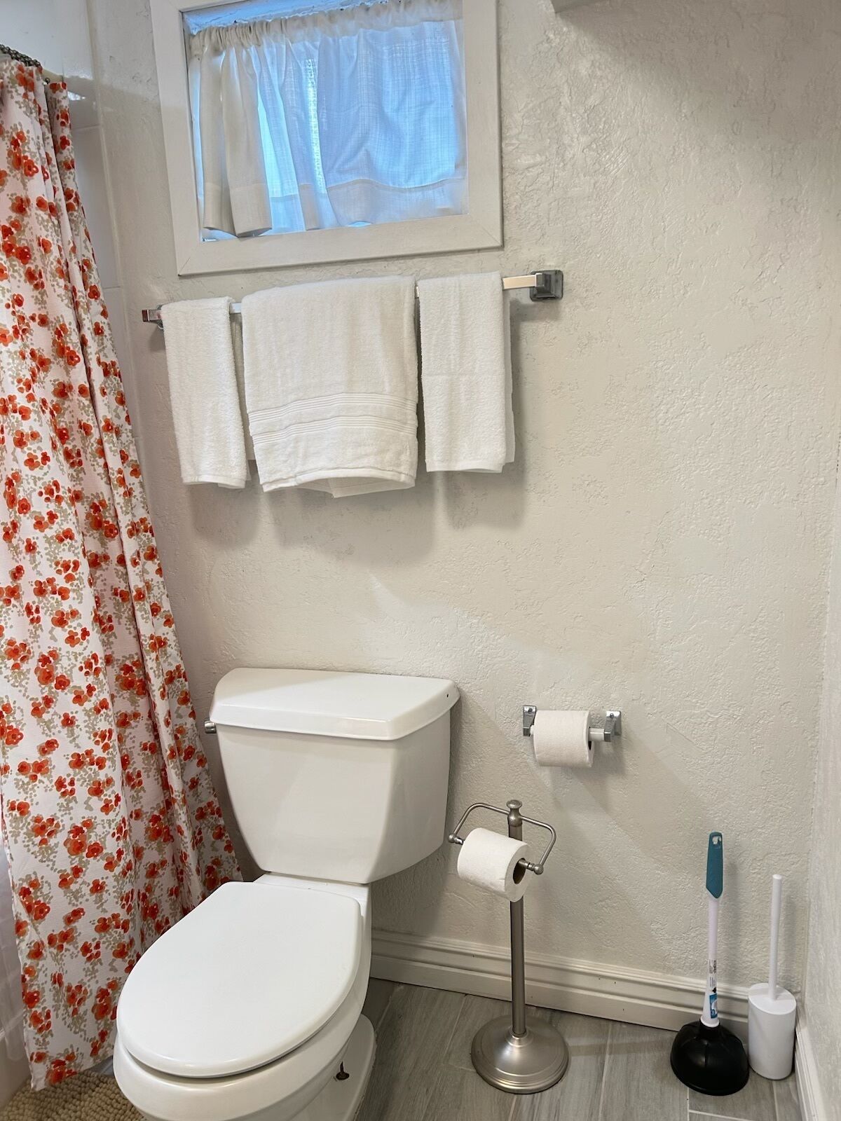 A bathroom with a toilet, a roll of toilet paper, a plunger, and a window with a curtain.