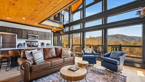 A spacious living area with a large brown leather sofa, two blue armchairs, and a wooden coffee table. A kitchen with dark cabinets and a large window is visible in the background. - Hill•of•a•View: Breathtaking Mountain Views & New Retreat (Sevierville)