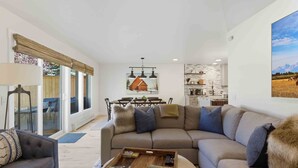 A modern living room with a sectional sofa, a coffee table with board games, a dining area with a table and chairs, and a kitchen with white cabinets and a stone backsplash. - The Timberline House | Modern Mountain Comfort | Walk to Town & Near Lifts (Ketchum)