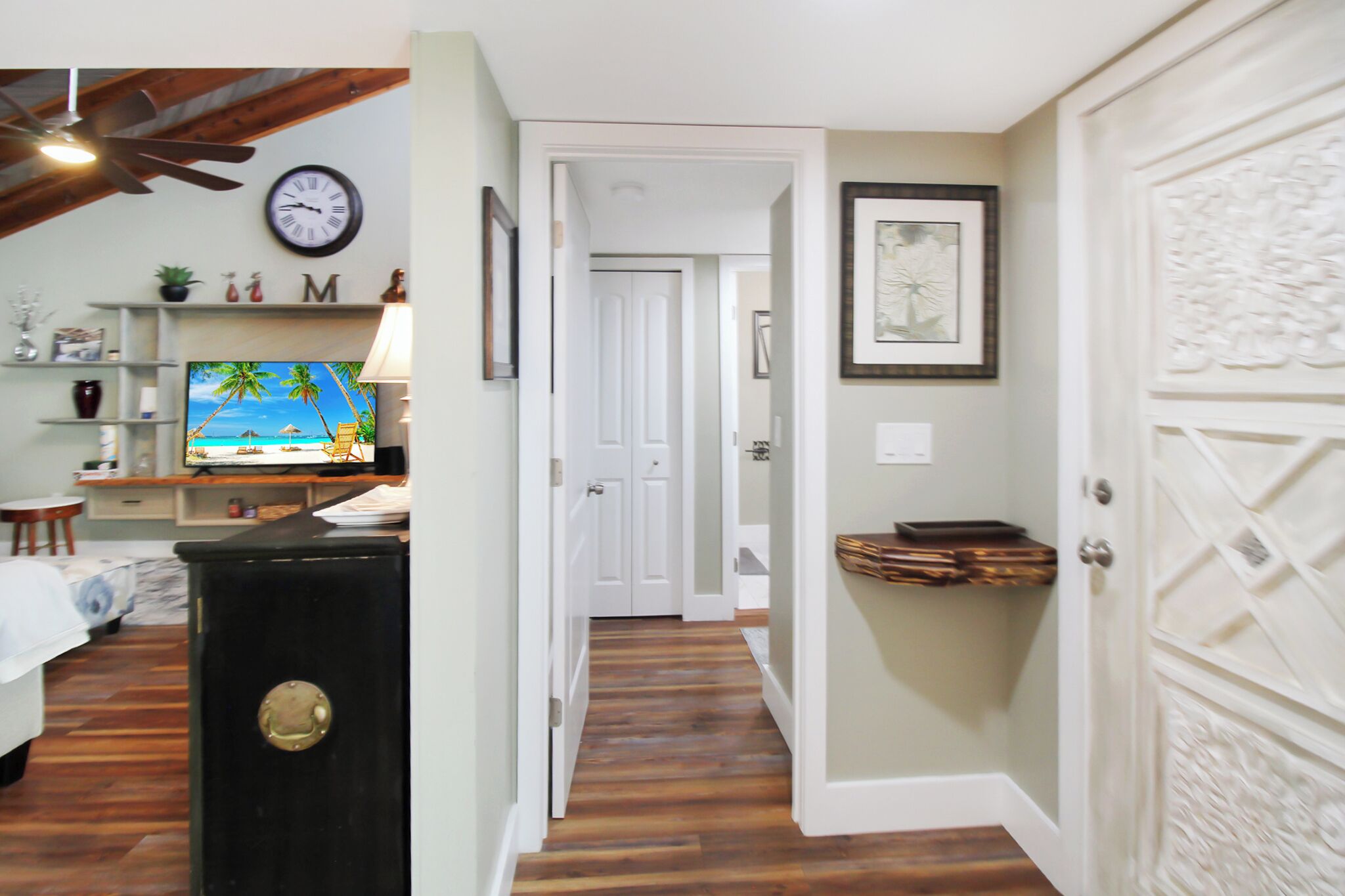 A hallway with wooden flooring, a television displaying a beach scene, a clock, and a door with decorative paneling.