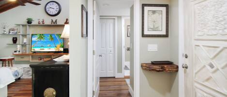 A hallway with wooden flooring, a television displaying a beach scene, a clock, and a door with decorative paneling.