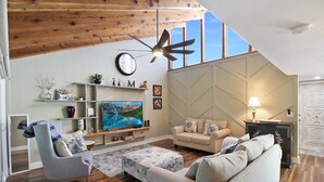 Un salon spacieux avec un plafond haut, des poutres apparentes, un grand téléviseur et un coin salon confortable avec un canapé et un fauteuil.