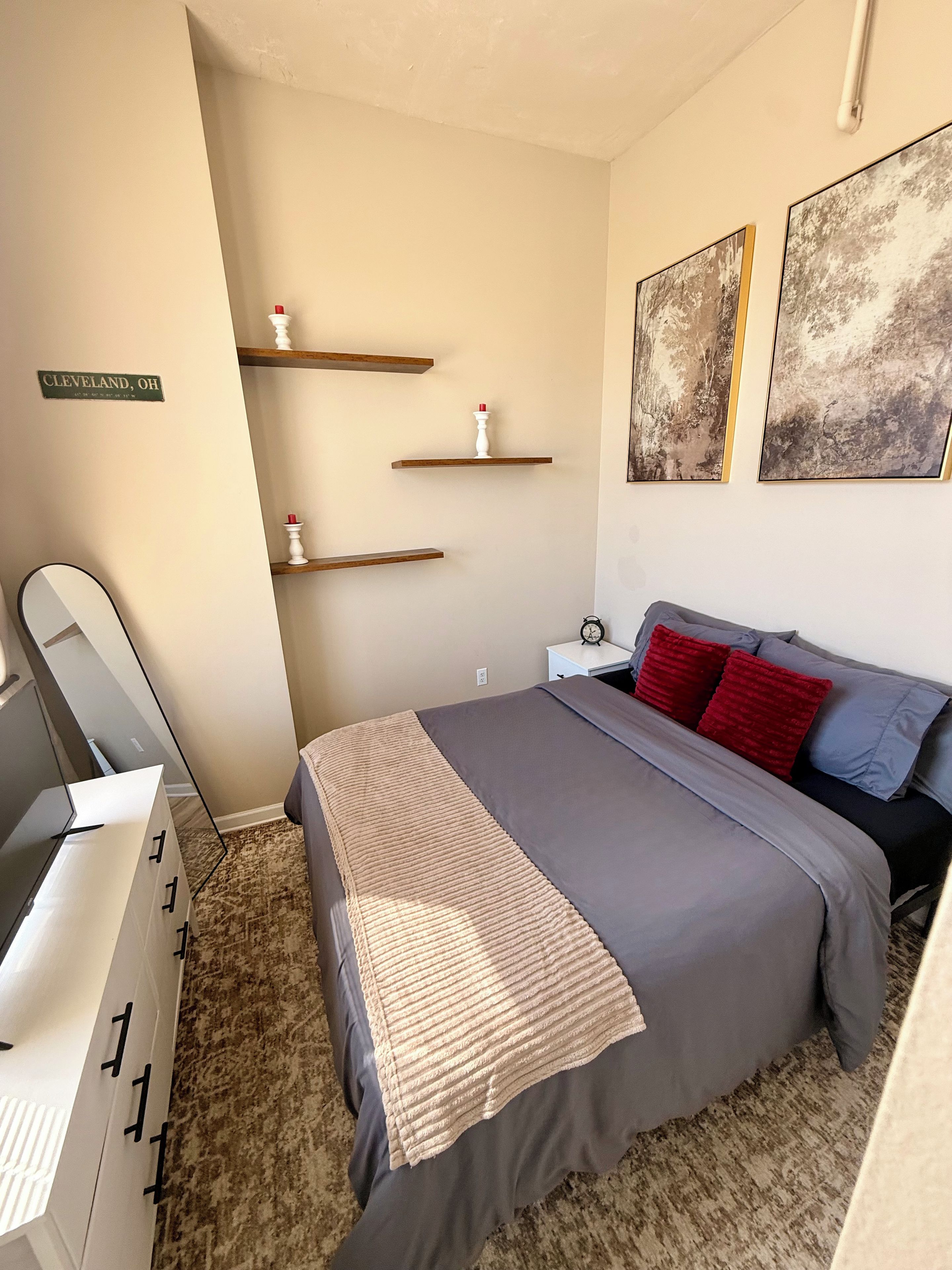 A bedroom with a bed, bedside table, nightstand, and a mirror. There are wall-mounted shelves with decorative items.