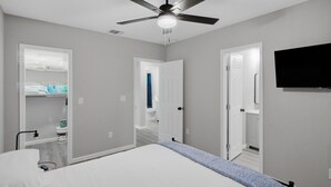 Une chambre avec un lit, un ventilateur de plafond, un téléviseur fixé au mur et un couloir menant à une salle de bain.