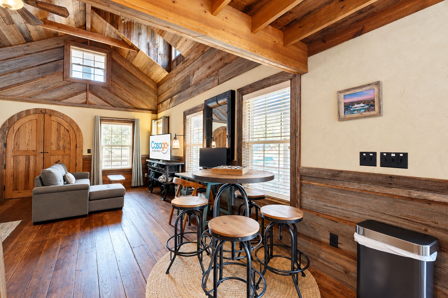A rustic interior with a wooden ceiling, a sofa, a TV, and a dining area with stools.