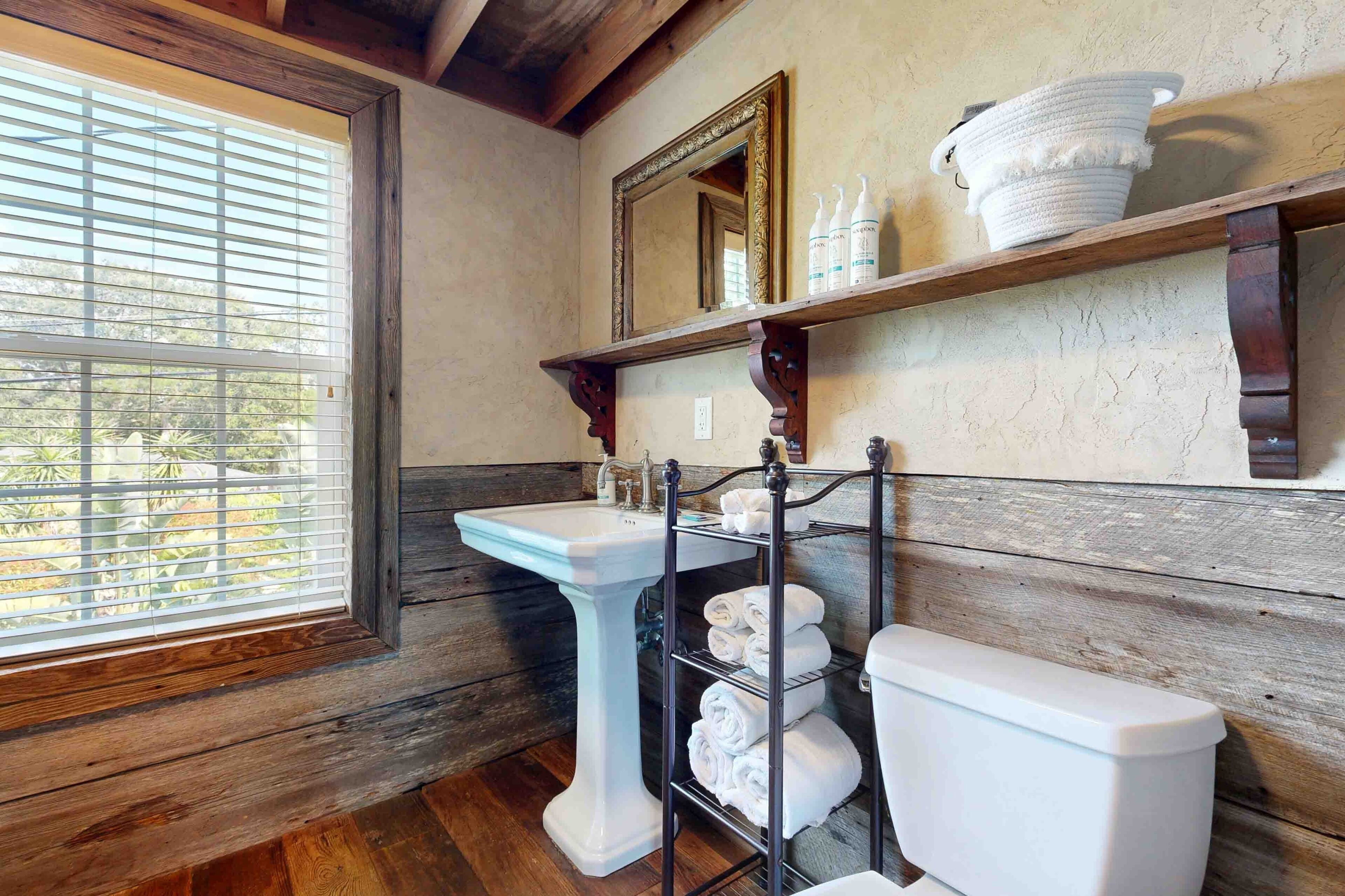 A bathroom with a wooden floor, a pedestal sink, a toilet, and a shelf with towels and toiletries.