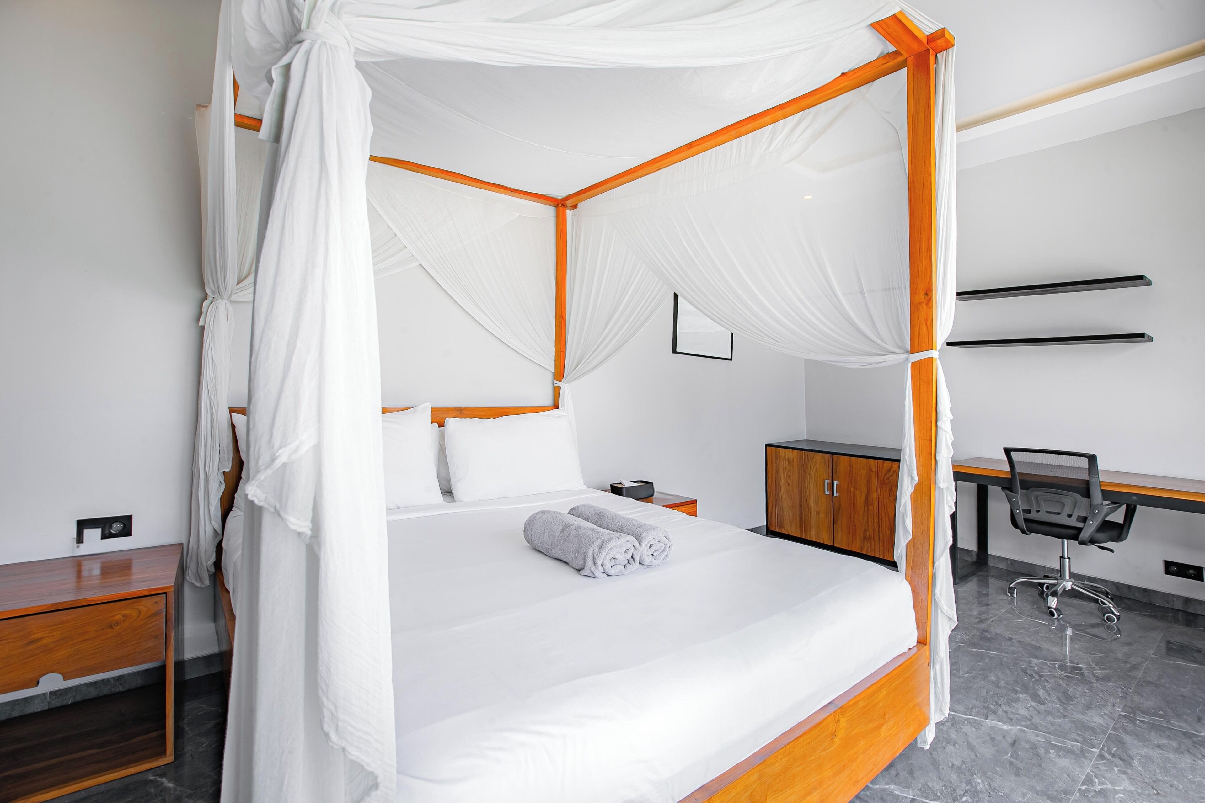A four-poster bed with white linens and a canopy, a wooden desk with a chair, and a nightstand with a lamp.