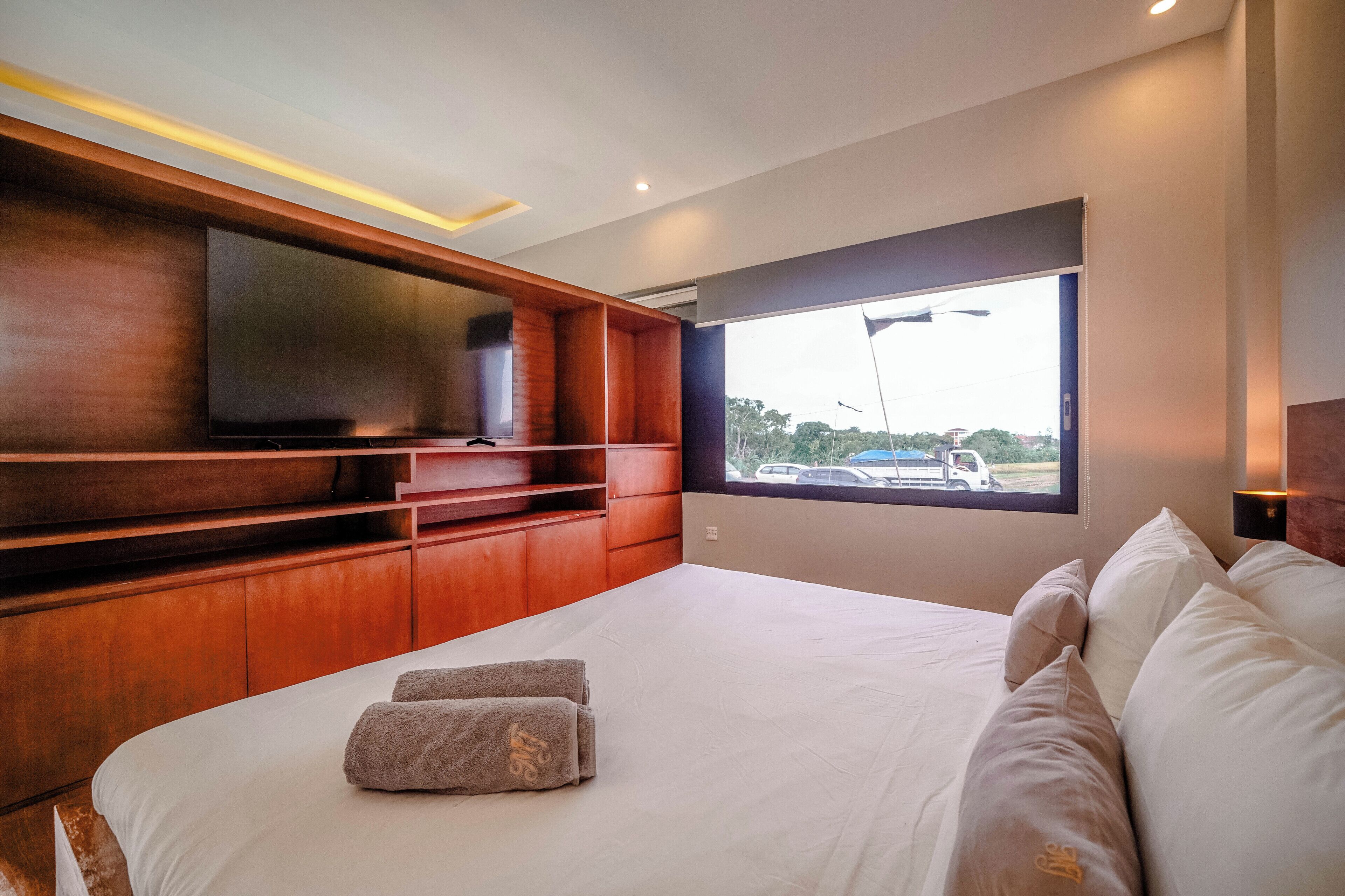 A modern hotel room with a large bed, a flat-screen TV mounted on a wooden panel, a window with a view of the outside, and a built-in shelf.