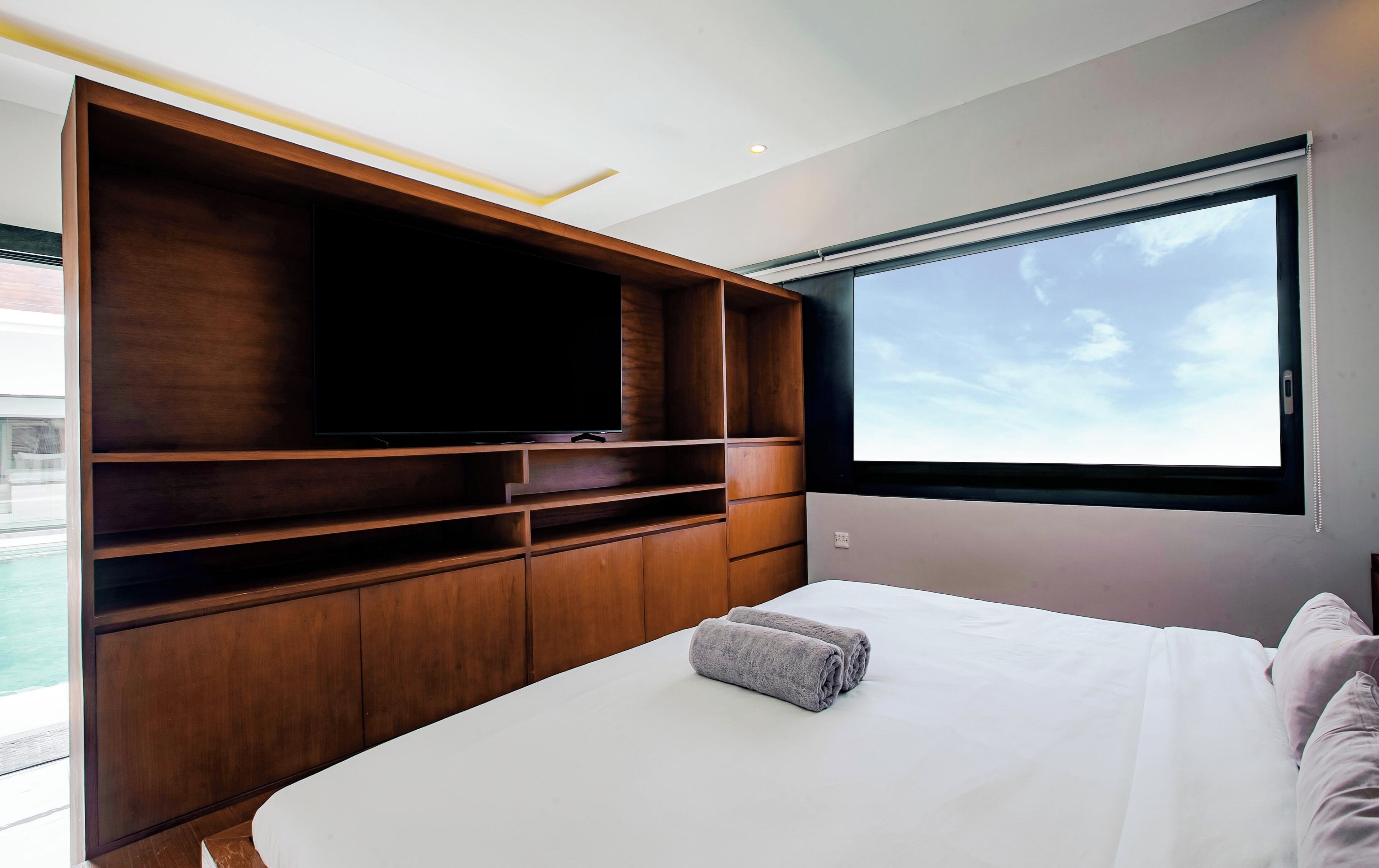 Un dormitorio con una televisión grande sobre una estantería de madera, una cama con ropa de cama blanca y un ventanal con vistas al cielo.