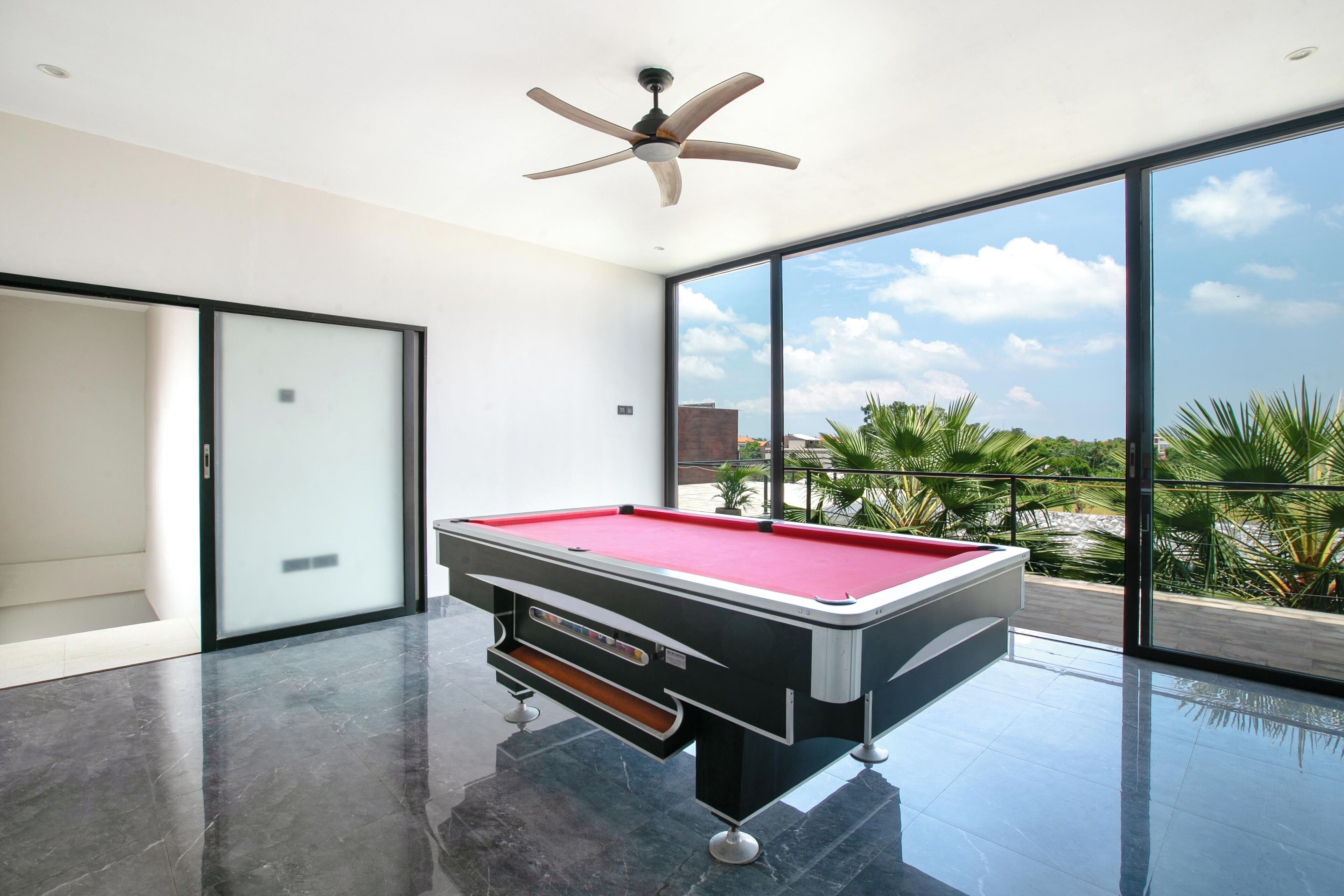 Une table de billard moderne pour intérieur, avec un drap de jeu rouge, un ventilateur de plafond et une grande fenêtre offrant une vue sur les palmiers et les bâtiments.