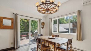 A dining area with a wooden table, chairs, a chandelier, and a view of the outdoors through large windows. - New! Large Patio, Dogs Allowed, Great Location! (Whitefish)