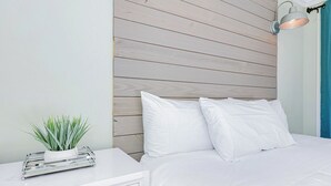 Een opgemaakt bed met witte lakens, een houten hoofdbord, een nachtkastje met een plant en een wandlamp.