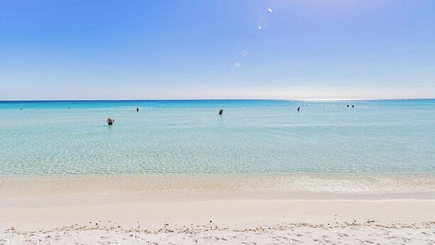 Een strand met helder turquoise water, een zandstrand en enkele mensen die zwemmen.