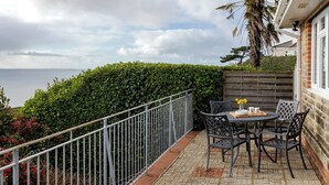 A patio with a table and chairs, a view of the sea, and a hedge.