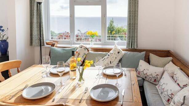 A dining table set for four with plates, cutlery, and wine glasses, a vase with yellow flowers, and a view of the sea through a window.