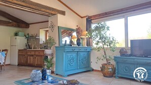A spacious living room with a large window, a kitchen area, and a blue cabinet. - Le Grenier d'Alfred - MOUZIEYS PANENS (MOUZIEYS PANENS)