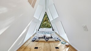 Une pièce étroite aux hauts plafonds, avec un lit placé au centre, juste sous une lucarne triangulaire. Le sol est en bois et la lucarne offre une vue sur une forêt.