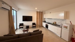 A modern living room with a sofa and chairs, a flat-screen TV mounted on the wall, and a kitchen area with white cabinets and a refrigerator. - Joli Beach House - Spacious Ground Floor T2 Apartment (Sal)