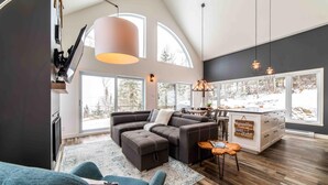 A spacious living area with a large window, a dark-colored sofa, a wooden dining table, and a kitchen island. - Petite Ourse 61: Hot Tub, Views &  Private Trails (Les Éboulements)
