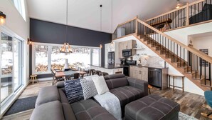 A modern living space with a large sofa, dining area, kitchen, and a staircase leading to an upper level. - Petite Ourse 61: Hot Tub, Views &  Private Trails (Les Éboulements)