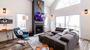 A modern living room with a fireplace, a television, and a large window offering a view of the outdoors. - Petite Ourse 61: Hot Tub, Views &  Private Trails (Les Éboulements)