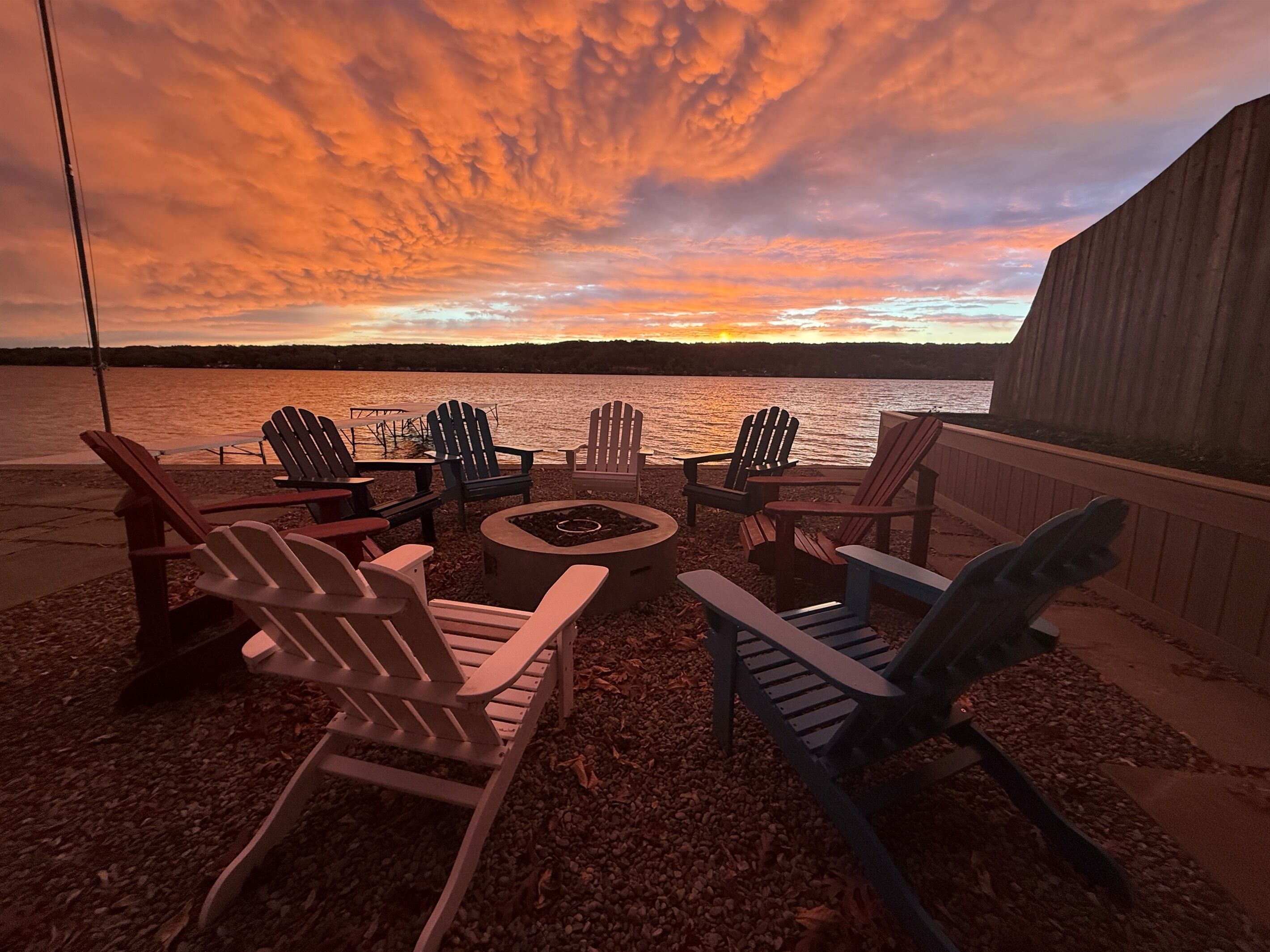 A lakeside seating area with Adirondack chairs and a fire pit, overlooking a sunset.
